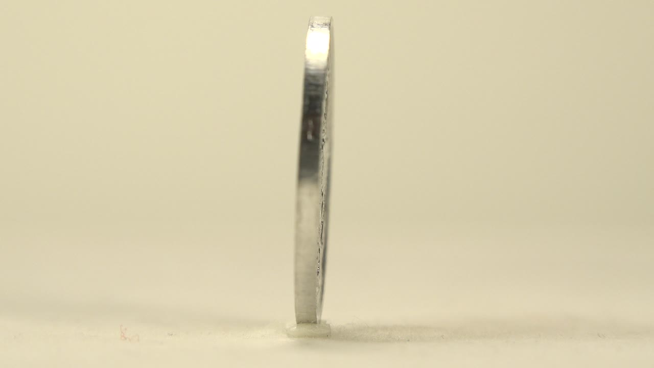The silver coin is nearly edge-on at the start of the clip, standing upright on a pale tabletop with soft warm background blur and no live people visible.