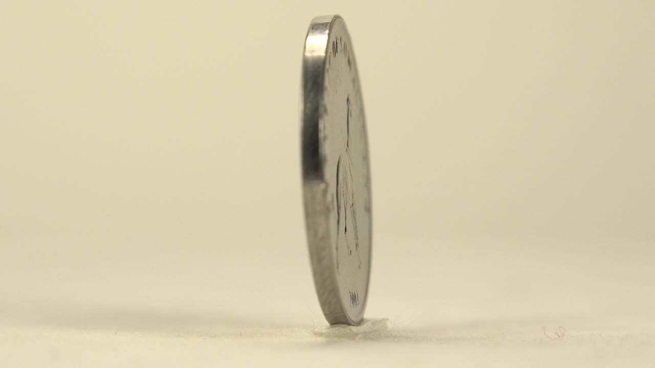 One second in, the upright coin remains mostly edge-on, with a small reflective face edge visible against the plain warm background and no visible people.