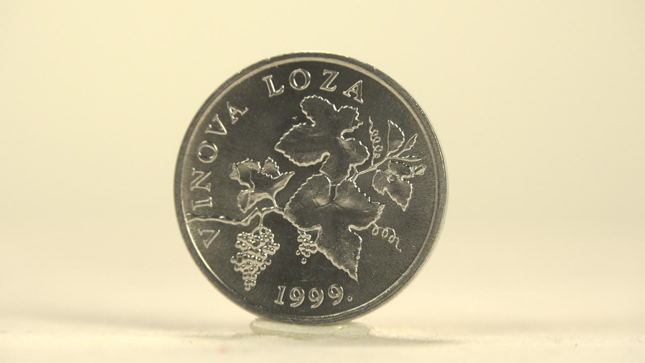 The silver coin remains upright and nearly face-on, with raised leaf details and curved lettering visible on a plain pale tabletop, with no visible people.