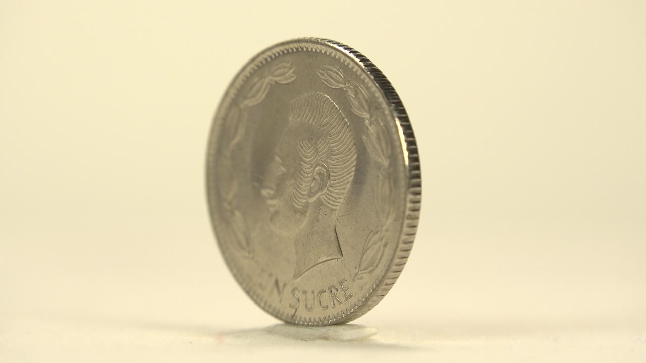 The coin remains upright on the pale tabletop, with the portrait side facing the camera more directly and no visible people.