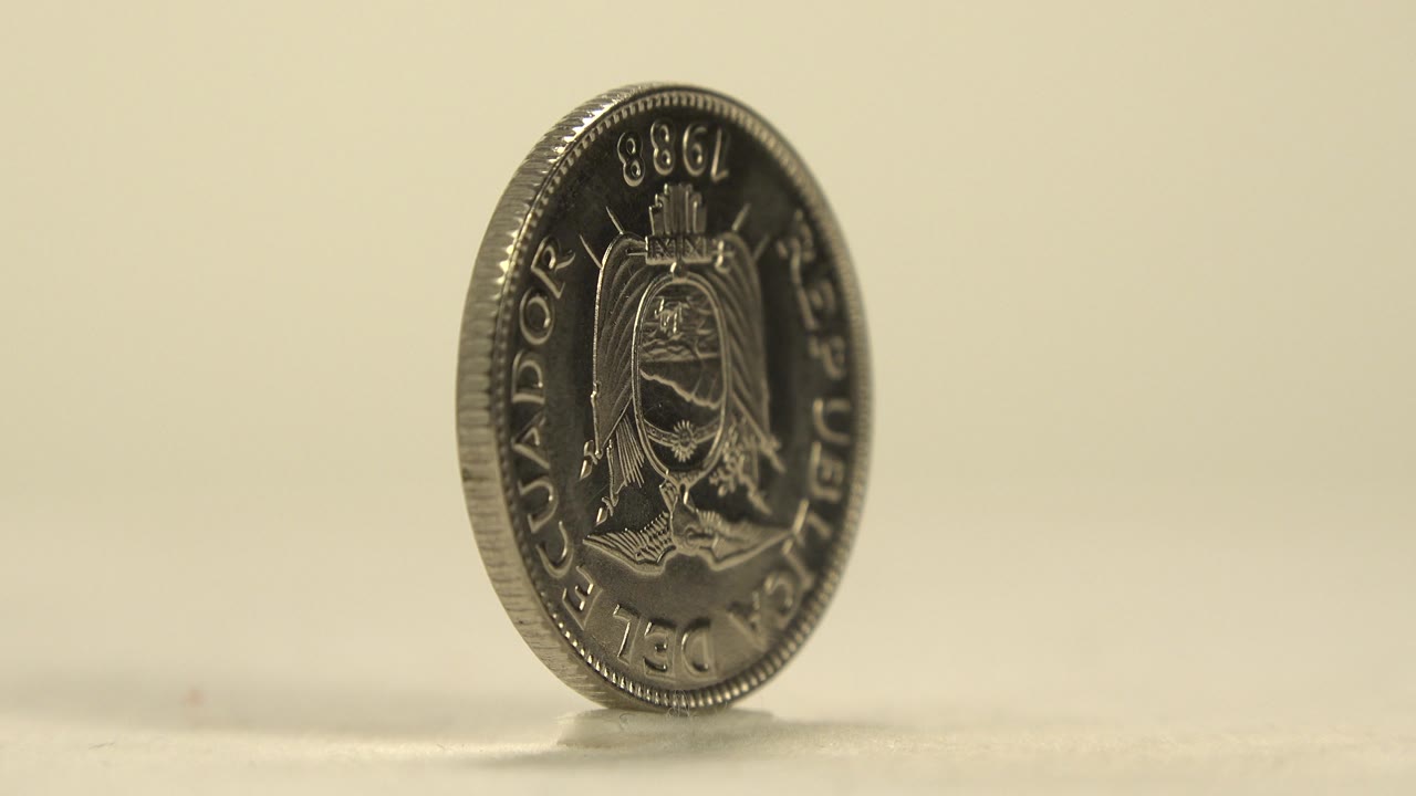 The coin rotates to reveal a darker crest side with raised lettering and a small shadow on the pale tabletop, with no visible people.