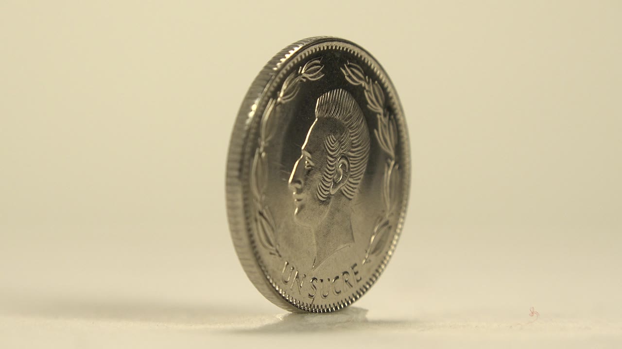 A silver coin stands nearly upright on a pale tabletop, showing a dark portrait side in profile with warm cream background and no visible people.
