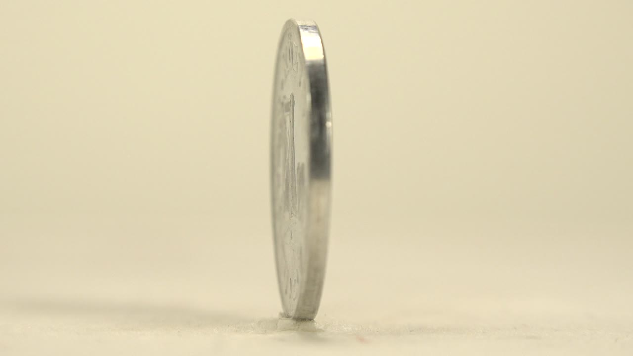 The coin turns almost edge-on, appearing as a narrow vertical silver strip on the pale surface, with no visible people.
