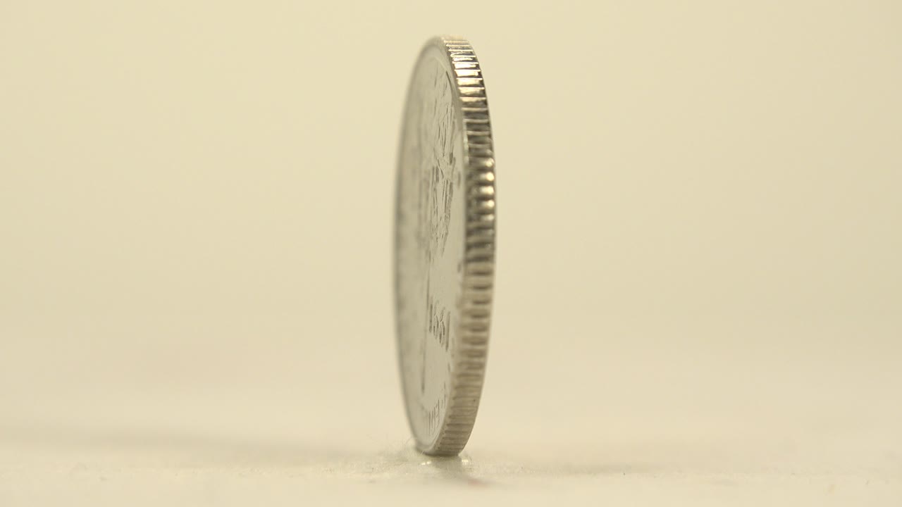 An upright silver-colored coin is shown mostly edge-on on a pale platform with a soft shadow and no visible people.