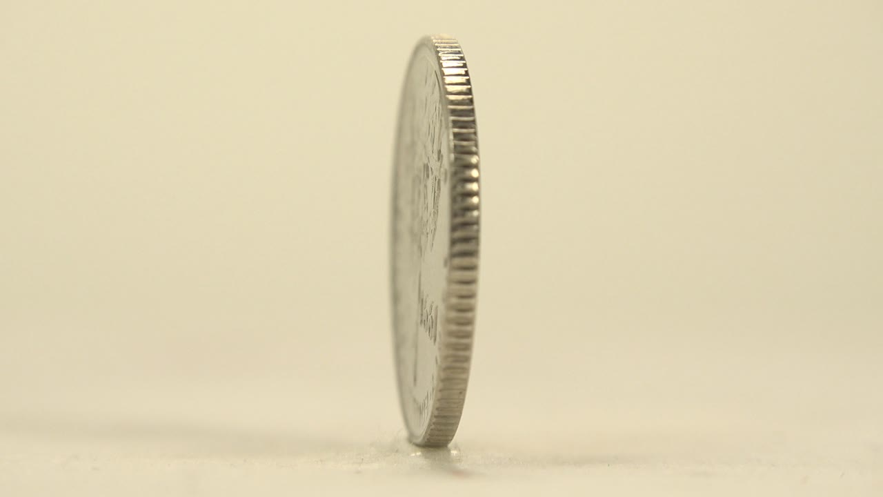 The coin stays upright in a narrow edge view, with the ridged rim catching warm light on the pale tabletop and no visible people.