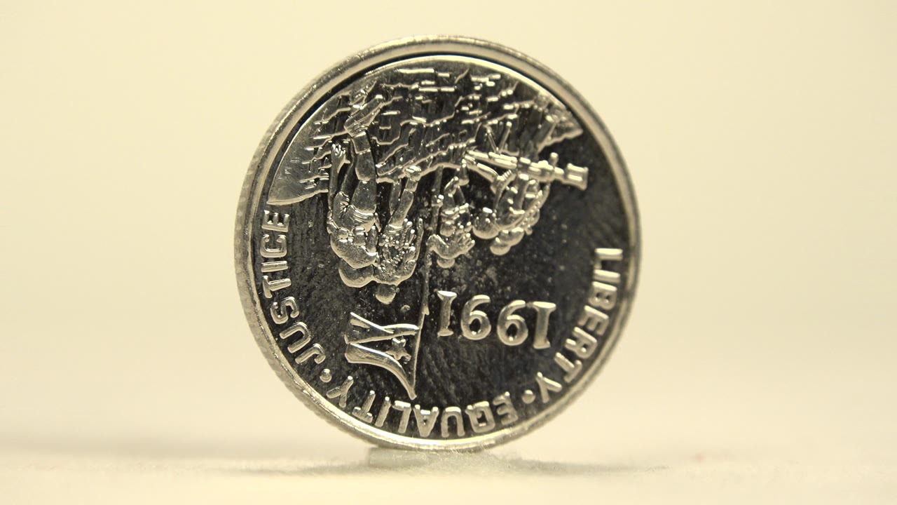 A silver coin stands upright on a white tabletop, showing engraved writing and the number 1997, with a pale background and no visible people.