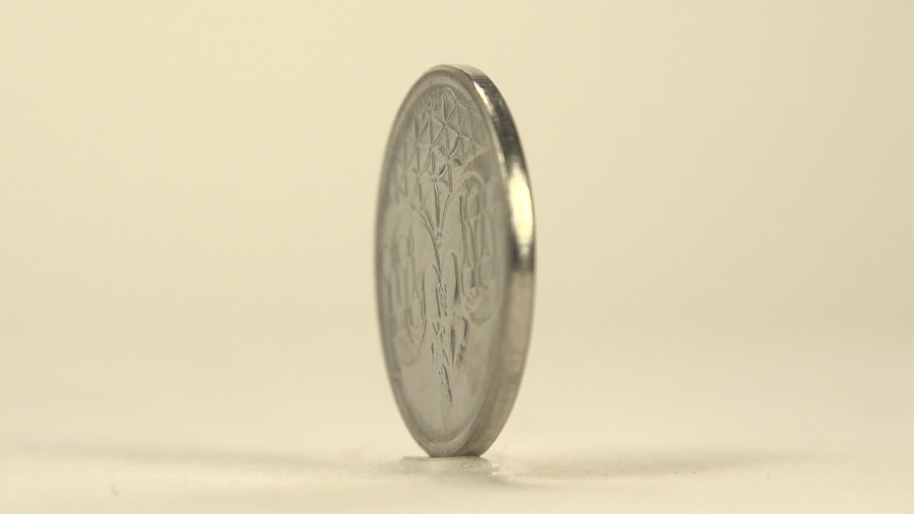 The coin remains upright on its edge with more of the reverse engraving visible, set low in the frame on the pale tabletop, no visible people.