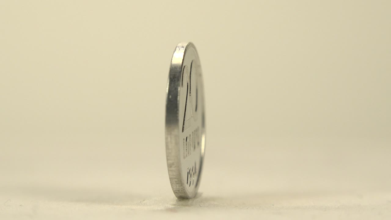 A silver coin is seen nearly side-on as it stands on its edge on a cream tabletop, with soft highlights and no visible people.