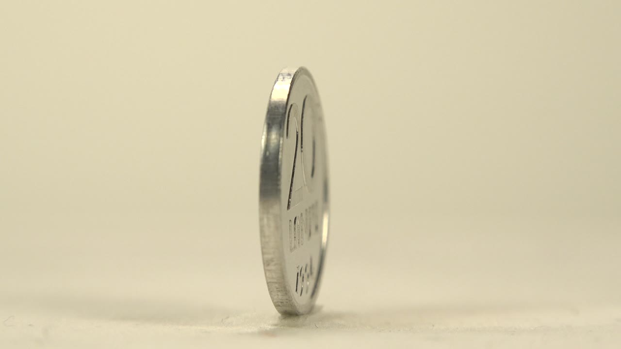The coin remains almost edge-on, slightly shifted in rotation, with a thin reflective rim against the plain cream background and no visible people.