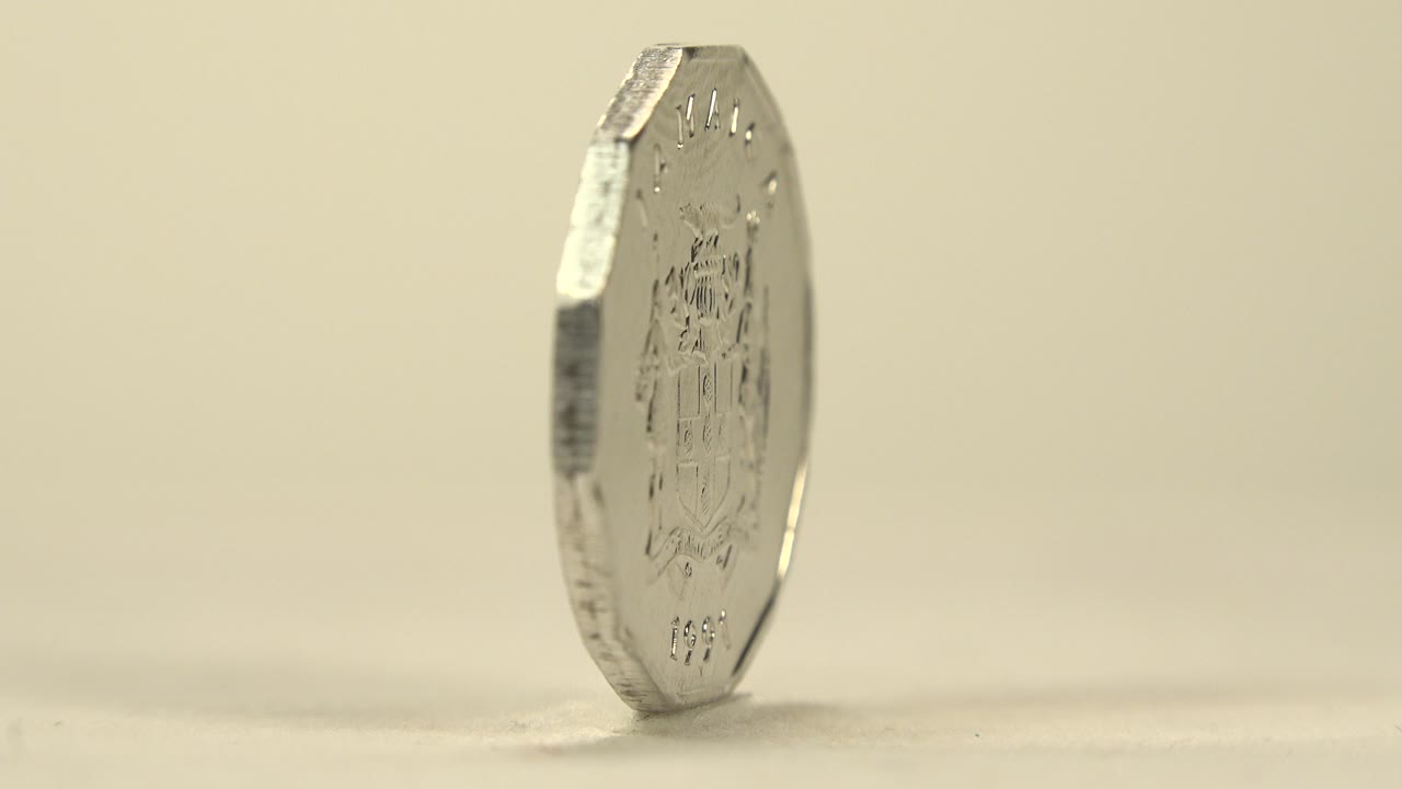 A many-sided Jamaican coin is seen mostly edge-on at the start of the turn, standing on a pale tabletop with soft cream background light and no visible people.