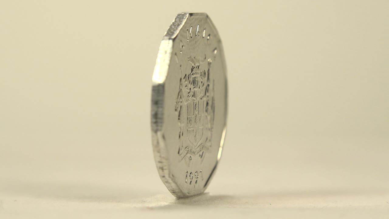 The coin remains nearly edge-on but slightly more of the face is visible, with faint raised markings catching soft light and no visible people.