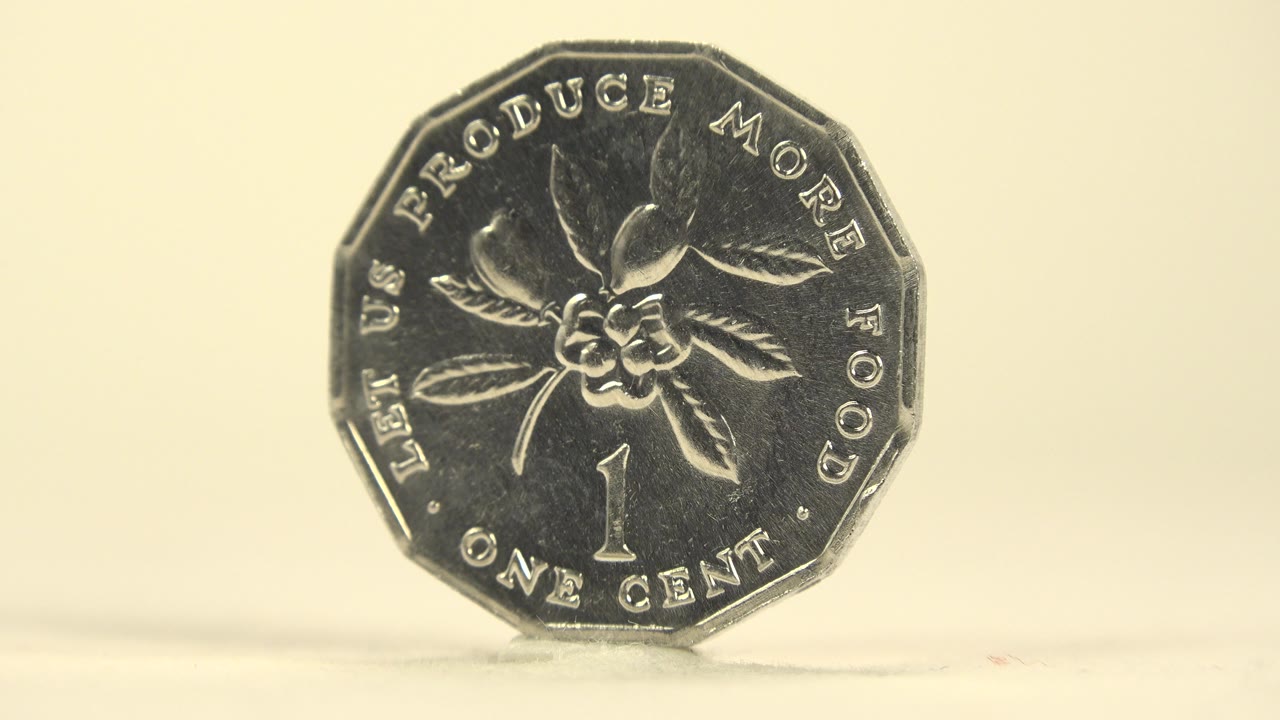 The Jamaican one cent coin remains nearly front-on, showing the flower engraving, raised edge, and reflective silver surface on a pale tabletop, with no visible people.