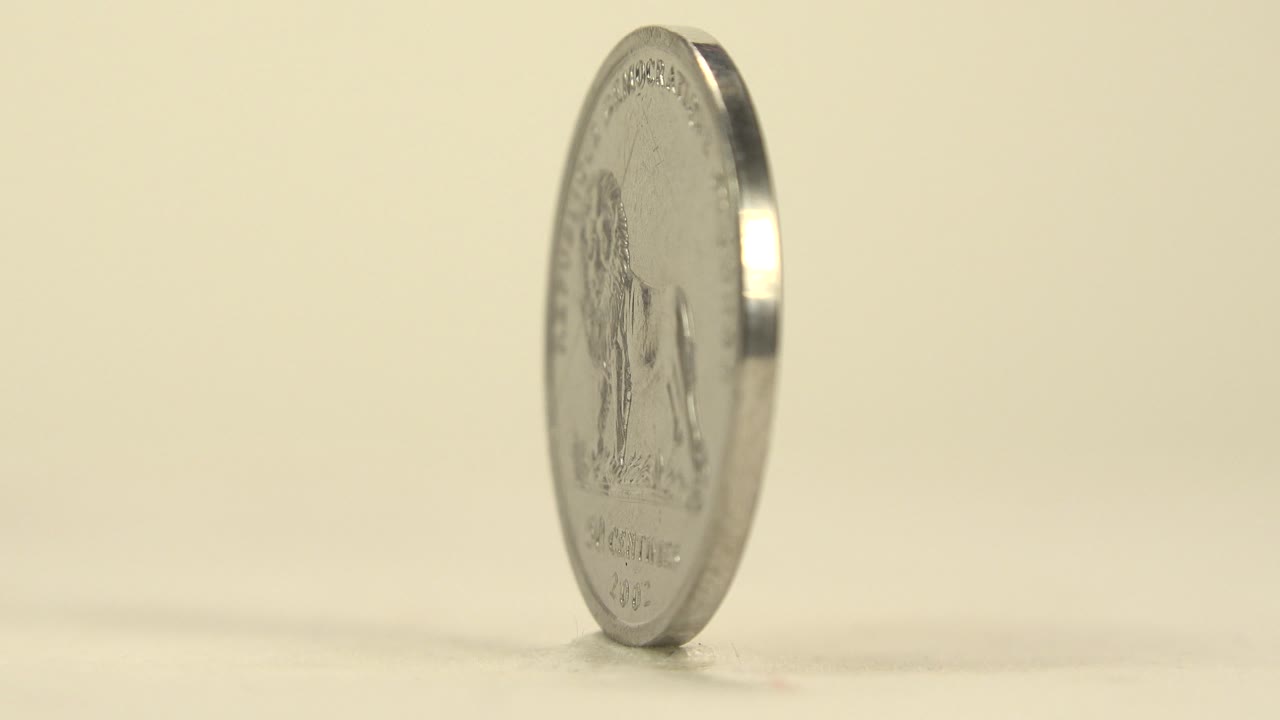 Silver coin remains upright on the tabletop, slightly turned so a partial engraved side catches soft light, with no visible people.