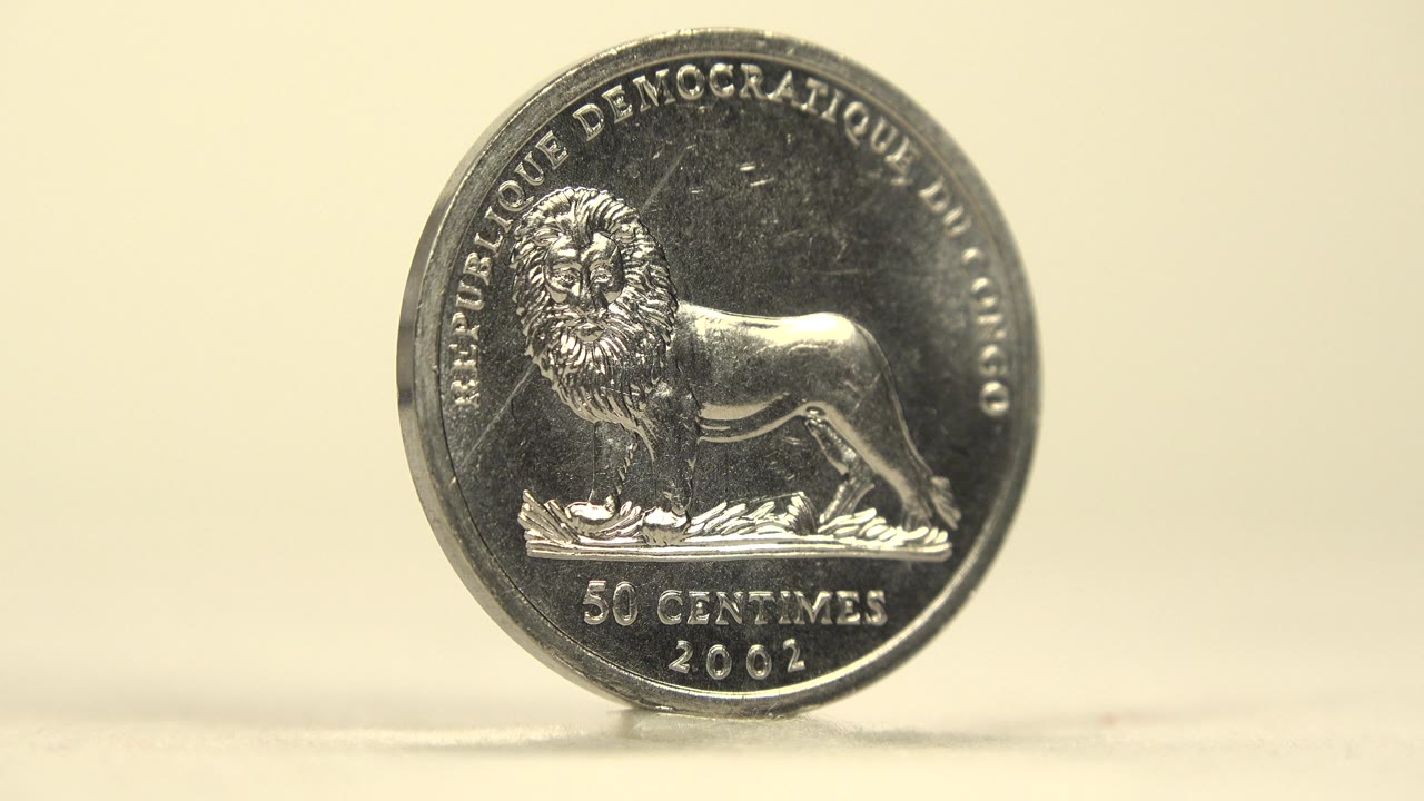 The coin remains upright with the lion face nearly front-on, its dark reflective metal catching highlights against the pale background and no visible people.