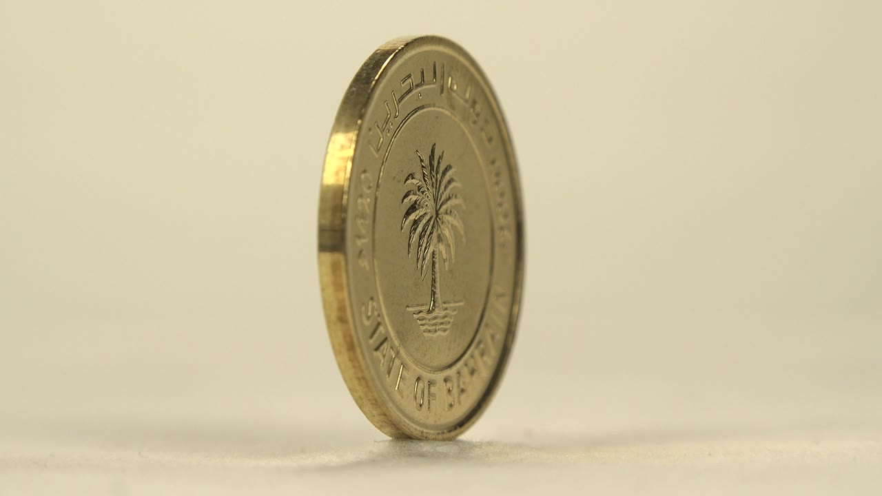 A gold-toned Bahrain coin stands on its edge at a left-facing angle on a pale surface, with the palm tree side partly visible and no visible people.