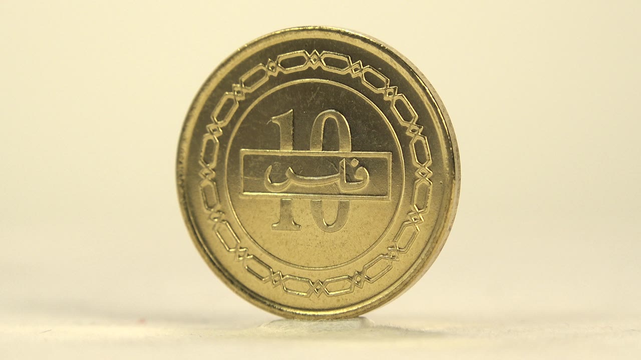 A gold Bahraini coin faces the camera on a pale surface, showing a large 10 marking, Arabic lettering, decorative border, warm yellow light, and no visible people.