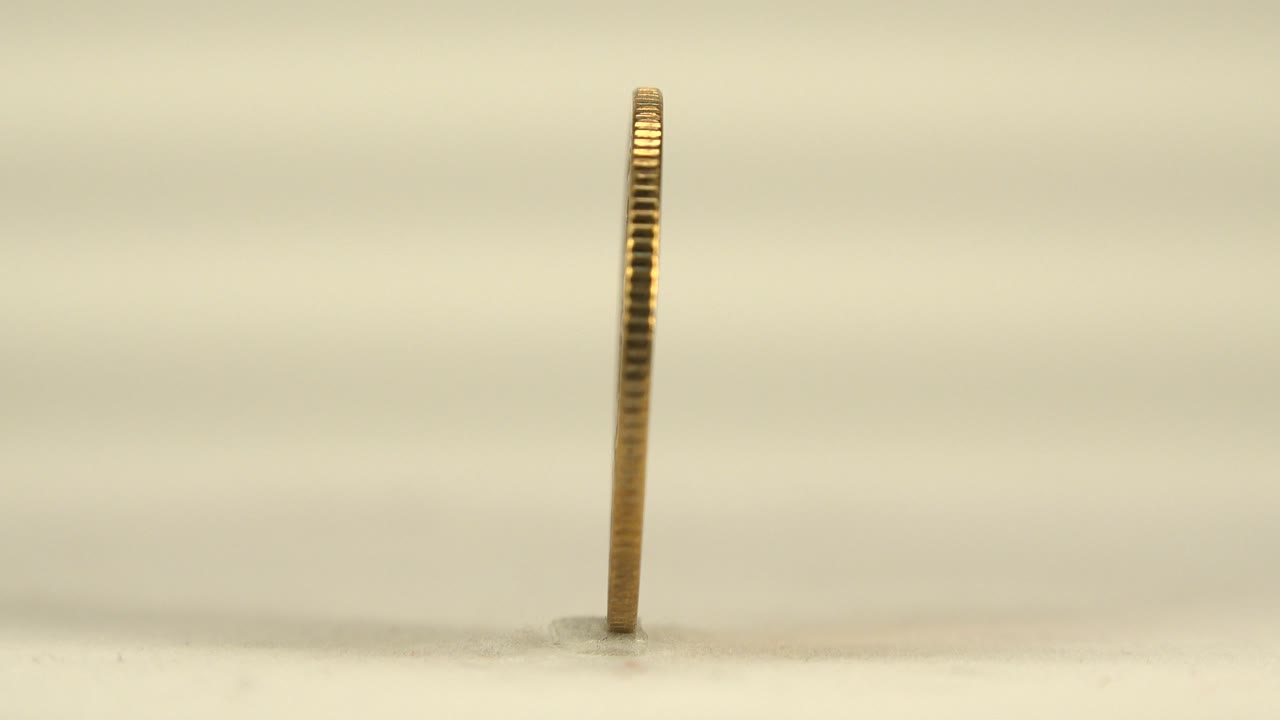 A bronze-gold coin is seen almost edge-on, standing upright on a pale reflective surface with a soft cream background and no visible people.