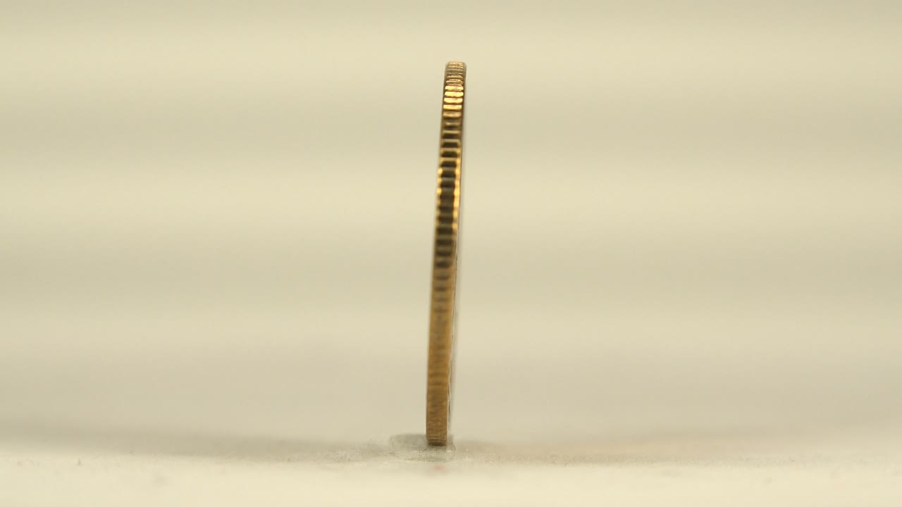 The coin remains nearly edge-on at center frame, with ridged metal edges catching warm light above the pale tabletop and no visible people.
