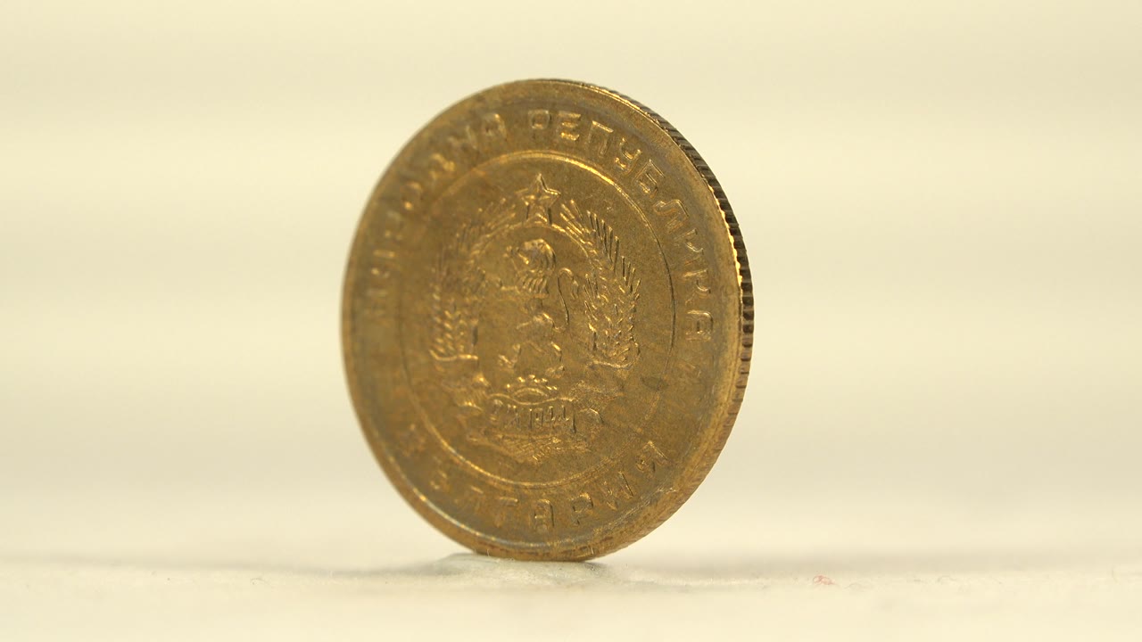 The coin has rotated toward a side angle, showing its thin ridged edge and partial face on the pale surface with no visible people.