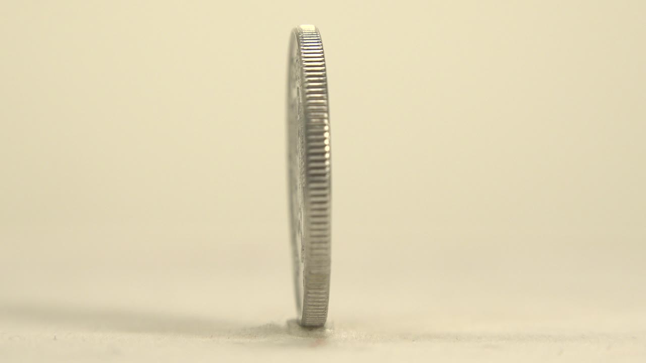 A silver coin is seen almost edge-on as it stands upright on a pale reflective plate, with a soft warm background and no visible people.
