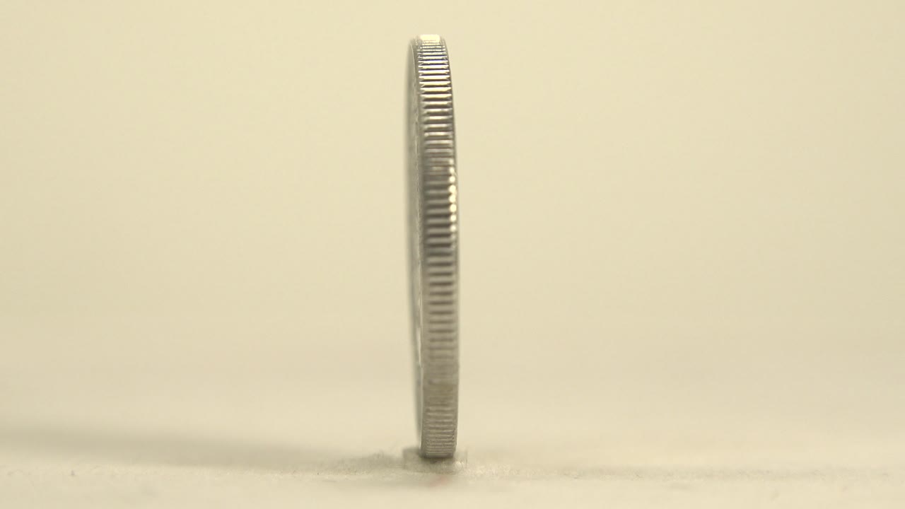 The coin remains nearly edge-on at plate level, showing its ridged rim, faint reflection, and soft shadow against a plain warm background with no visible people.