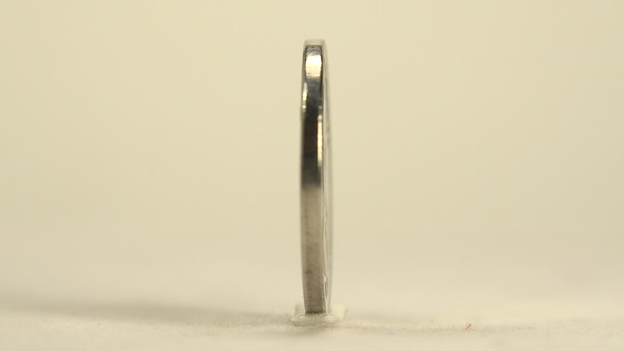 The coin appears nearly edge-on at the start of the clip, standing upright on a pale cream surface with warm light and no visible people.