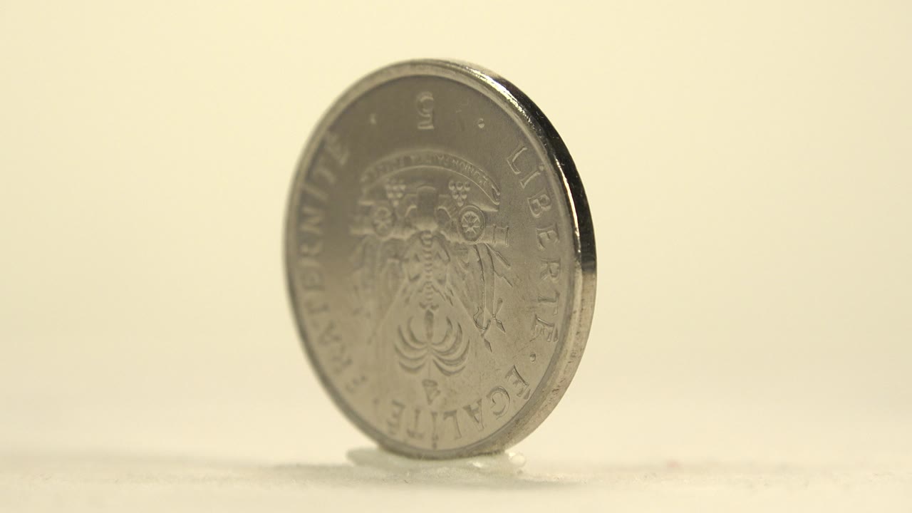 Silver-toned coin standing upright at an angle on a pale surface.