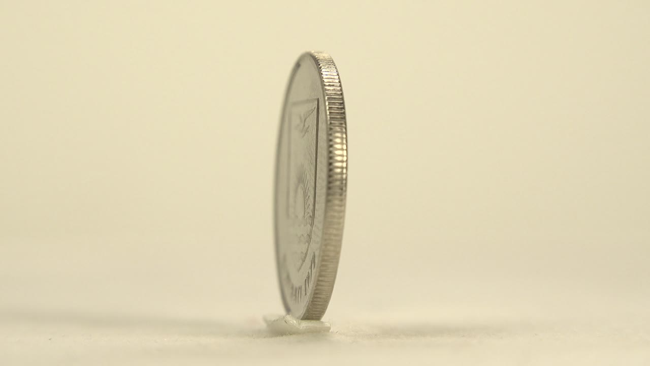 The upright coin remains near an edge-on angle, with its ridged rim catching light against the plain cream background and no visible people.