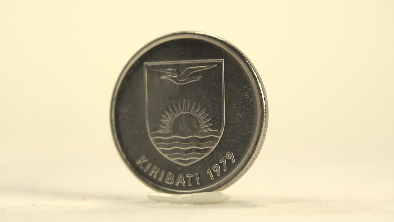 The coin remains upright on its small stand as the Kiribati crest side darkens slightly in the light, with a clean pale backdrop and no visible people.