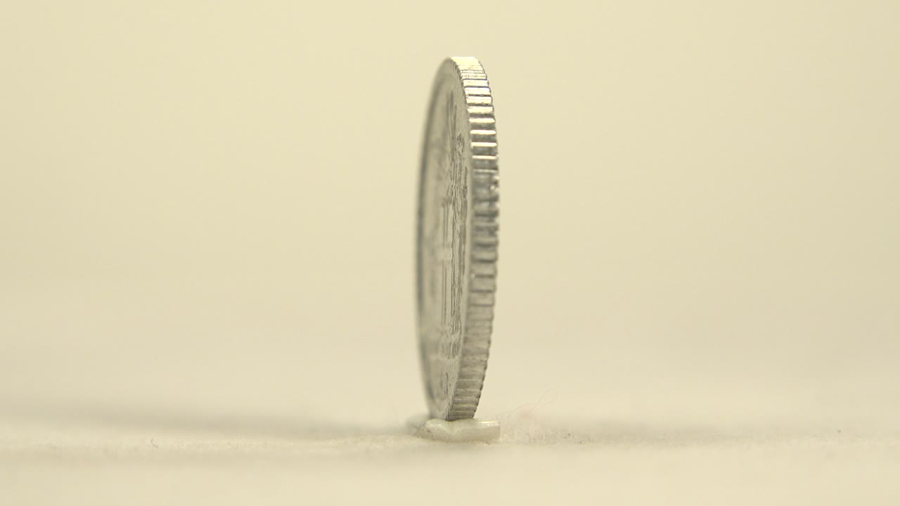 A silver coin stands nearly edge-on on a pale tabletop with soft warm light, shallow focus, and no visible people.