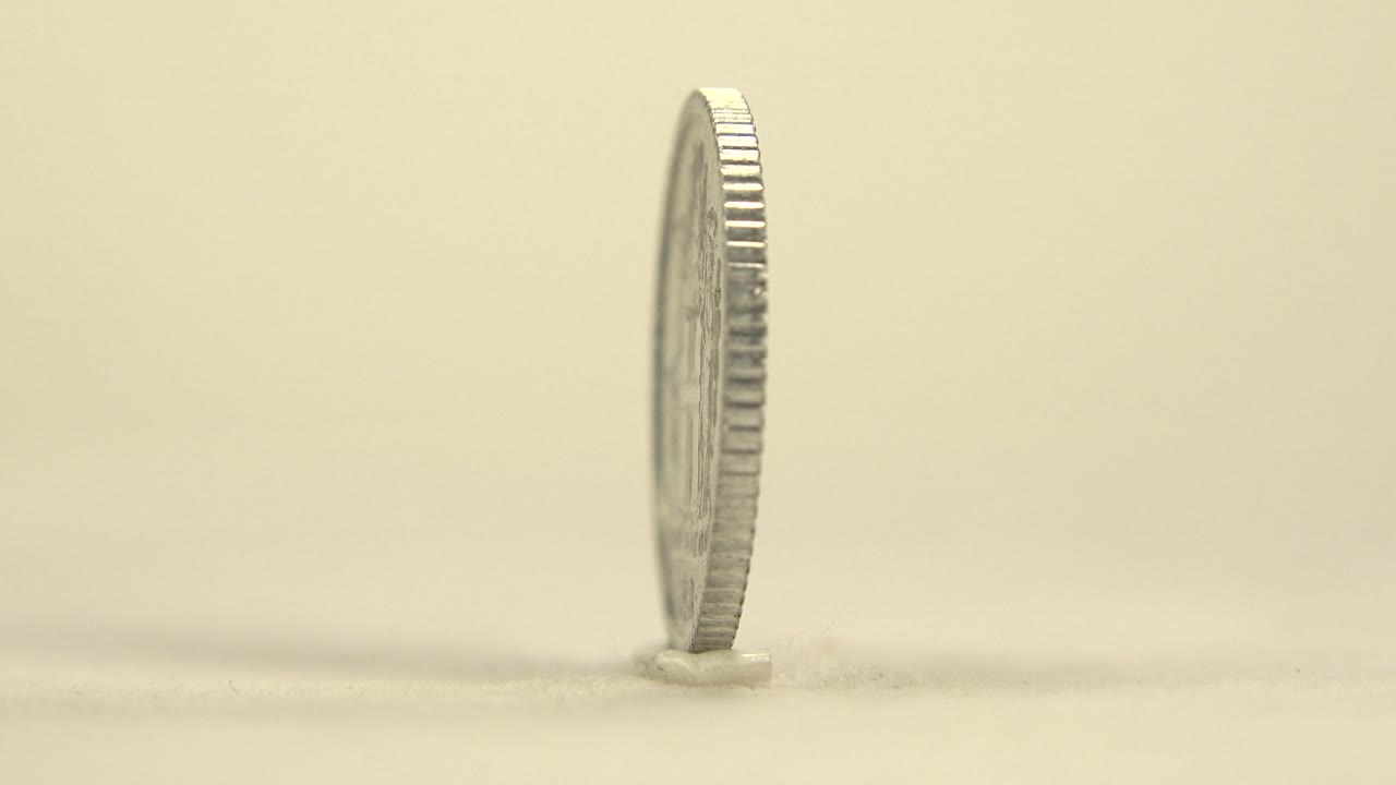 The coin remains almost vertical and edge-on, with ridged metal catching light against the plain cream background and no visible people.