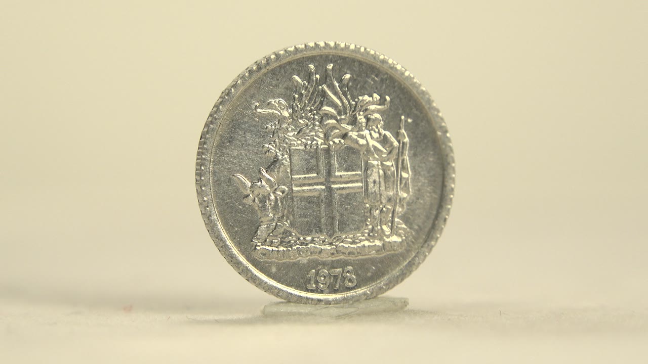 A silver coin stands upright on a pale surface, showing a crest design and the year 1978 against a warm cream background, with no visible people.