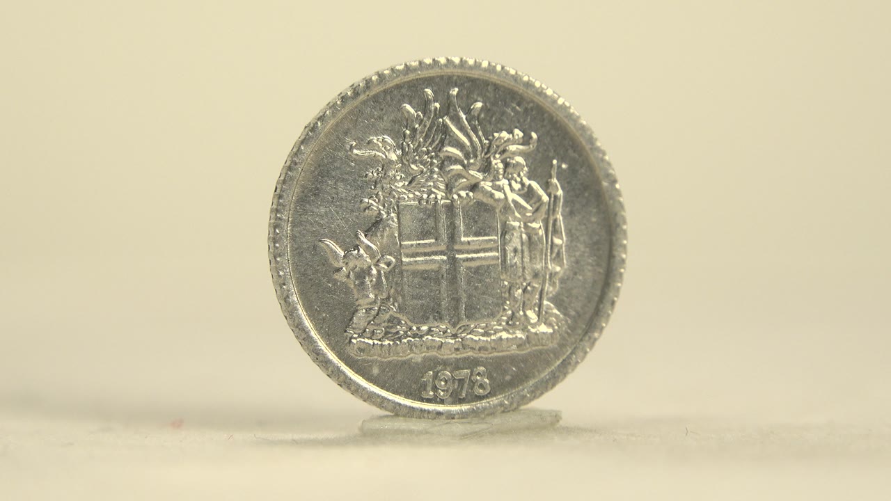 The coin remains upright with the crest side facing camera, its reflective surface catching soft light and no visible people in the frame.
