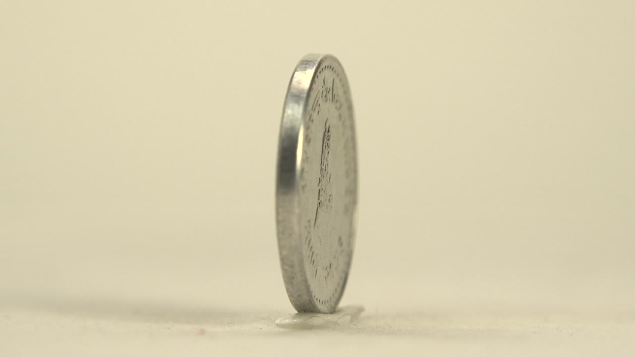 A narrow edge view of the silver coin stands on the pale surface, with the round face mostly turned away and no visible people.