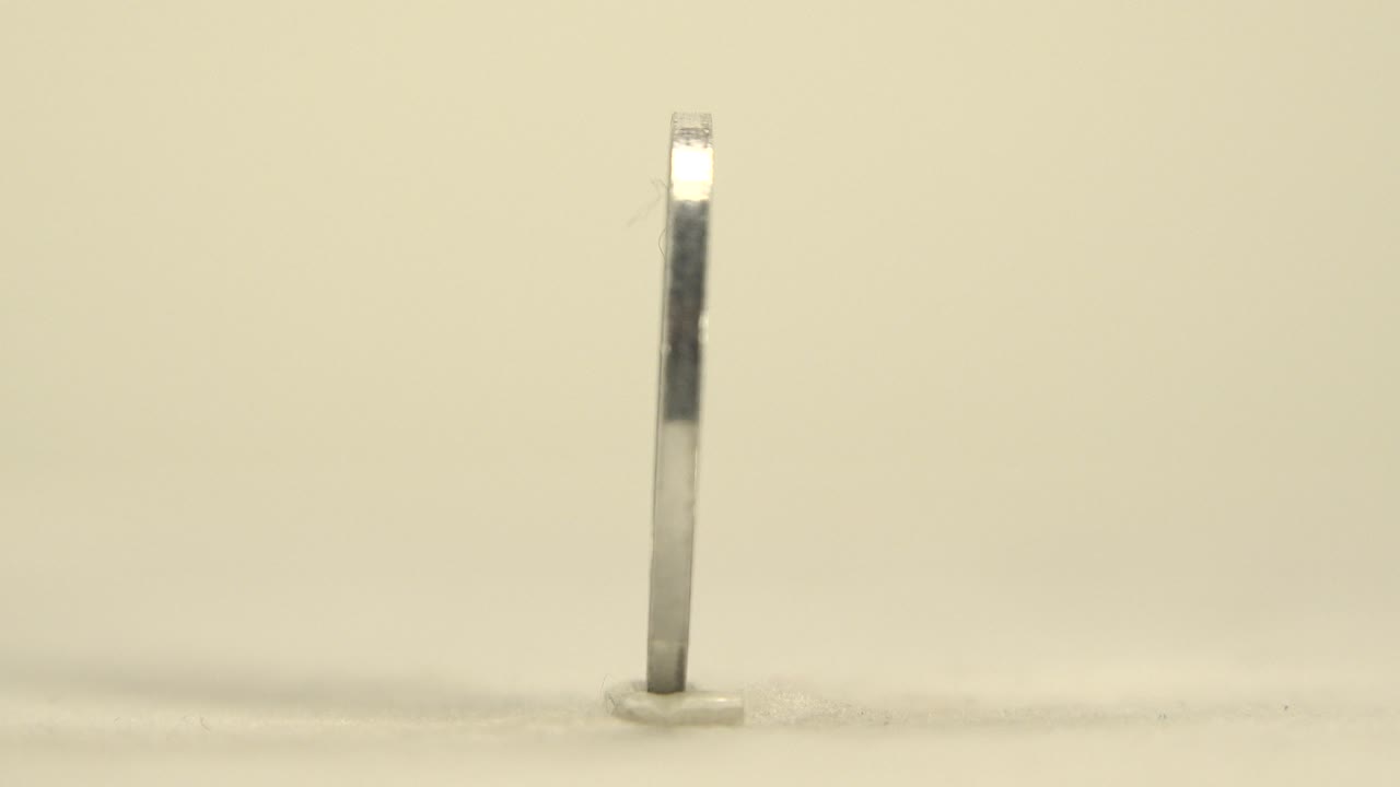 The coin remains upright and nearly edge-on near the center of the frame, with a small base contact point visible on the cream surface and no visible people.