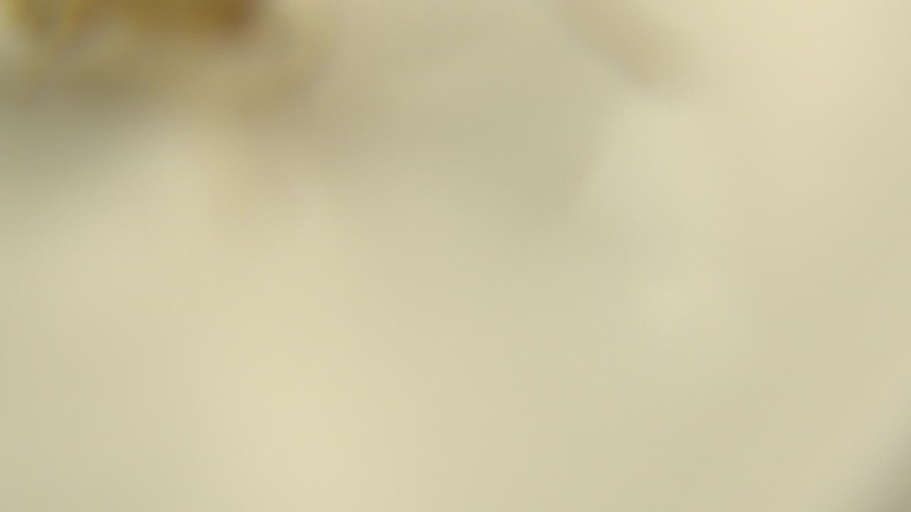 At 0:01, the close-up remains heavily out of focus, with pale yellow cream color and indistinct brown shapes near the top edge, no visible people.