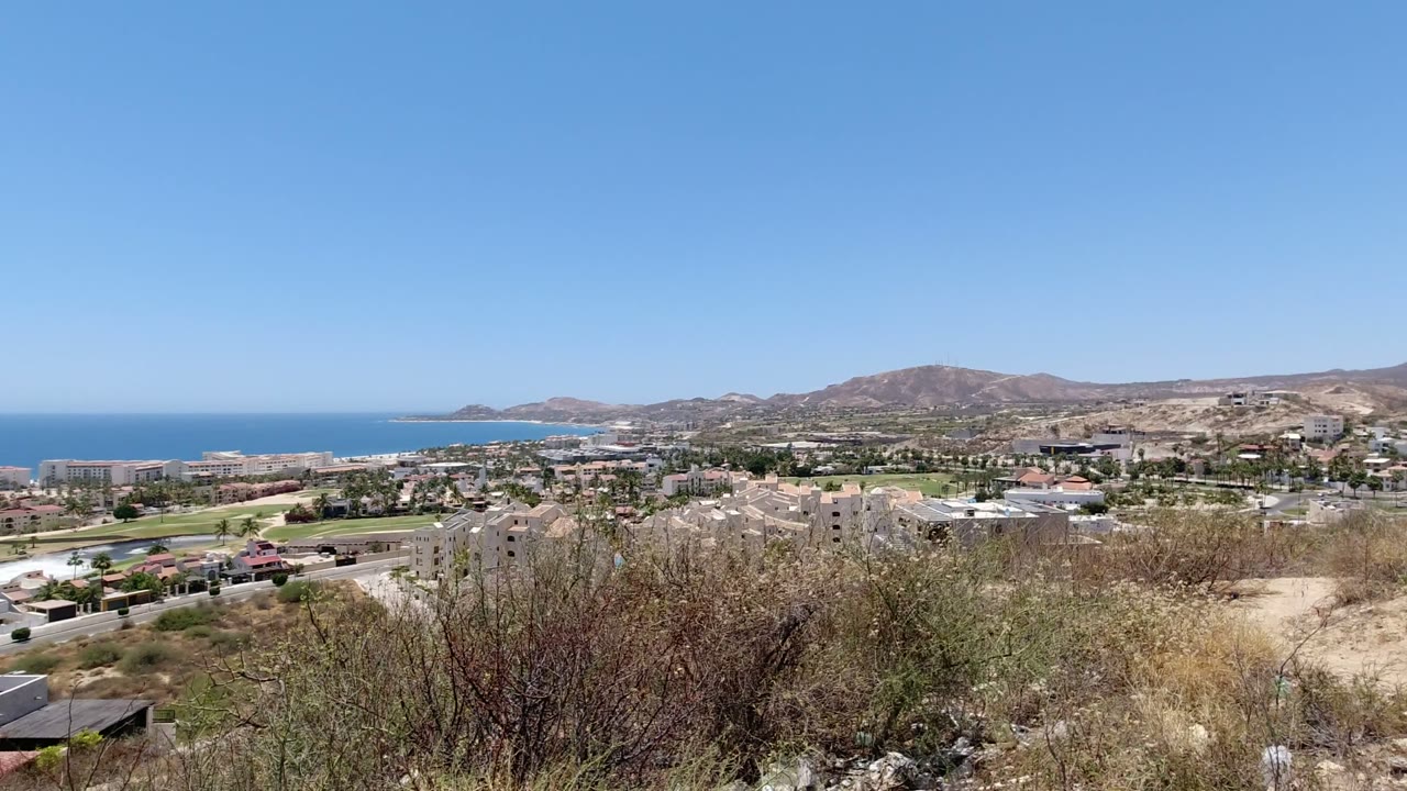 Wide elevated view over San Jose del Cabo with dry brush in the foreground, residential buildings below, brown hills behind, blue ocean at left, and no visible people.