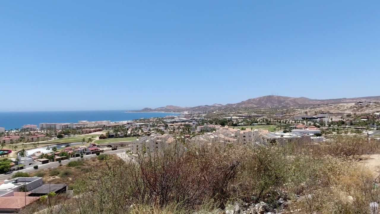 San Jose del Cabo appears from an elevated dry hillside, showing brush and rocks in front, low town buildings and roads in the middle distance, the ocean beyond, and no visible people.