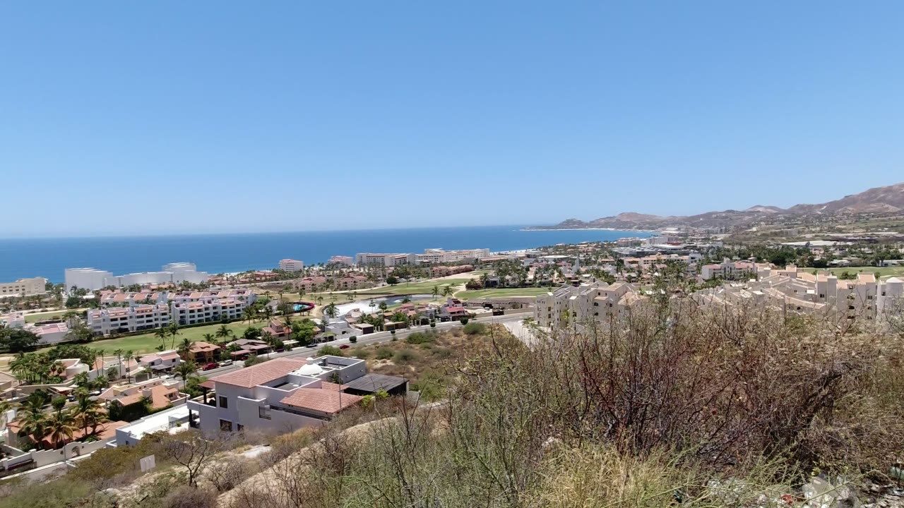 Hilltop view over coastal buildings, dry brush, blue sea, and clear sky.