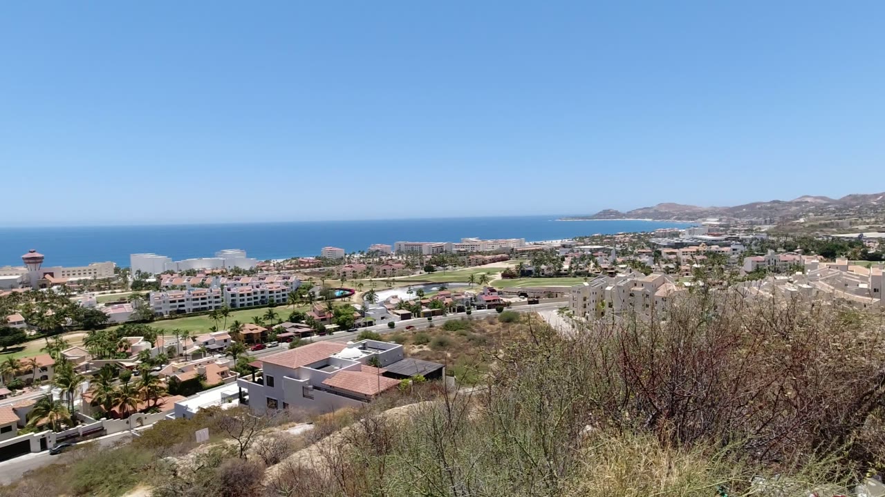 Early frame showing San Jose Del Cabo from above, with tan hillside plants across the lower edge, low-rise development in the middle, distant coastline, and no visible people.