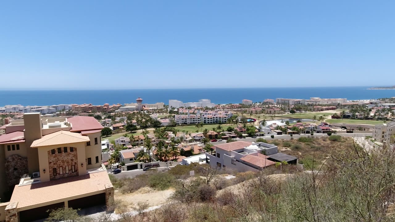 Elevated San Jose del Cabo view with dry hillside brush, tan homes, low buildings, blue ocean horizon, clear sky, and no visible people.