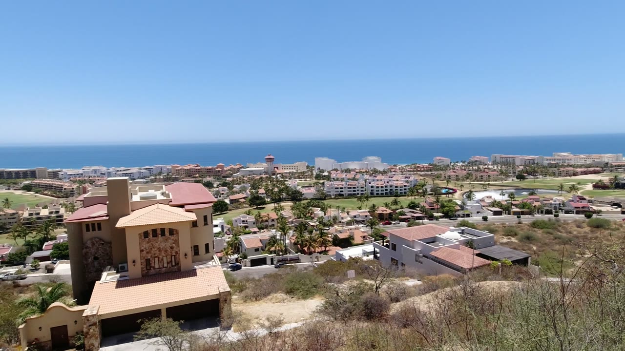 San Jose del Cabo frame centered on a tan hillside home, with residential streets, palms, resort-like blocks, blue sea, and no visible people.