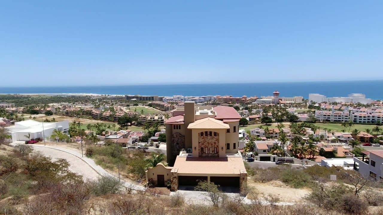 Elevated view over a tan house, dry hillside, coastal buildings, and blue ocean horizon.