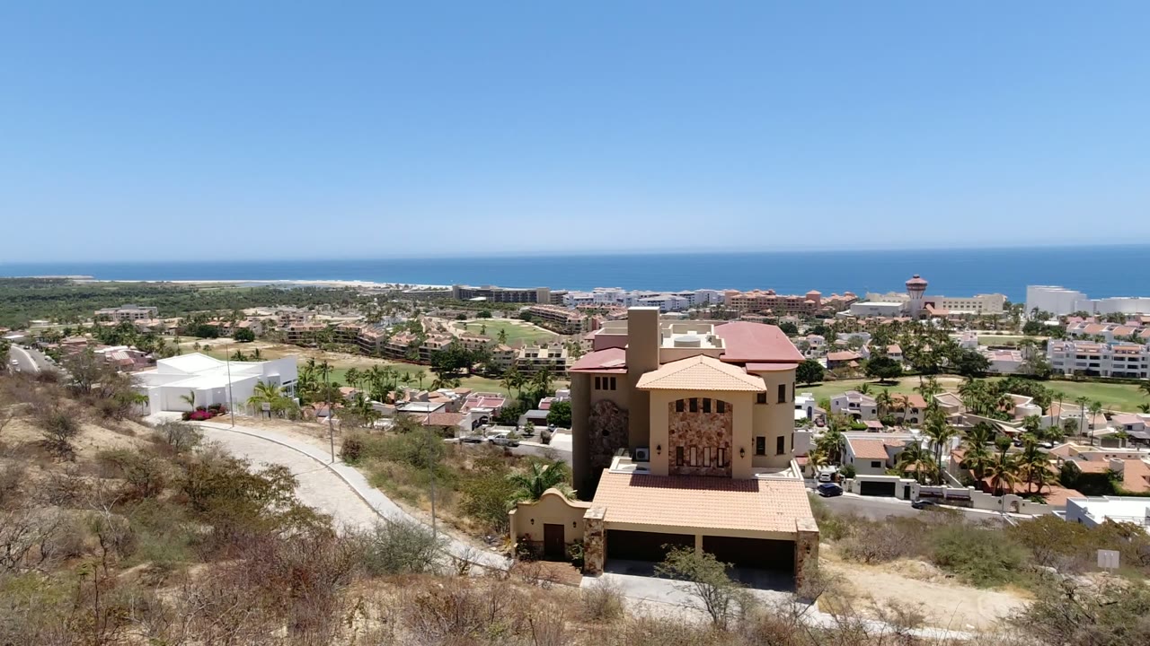 San Jose del Cabo hillside frame with the tan house more central, a curving road to the left, palms and low-rise buildings below, ocean horizon, and no visible people.