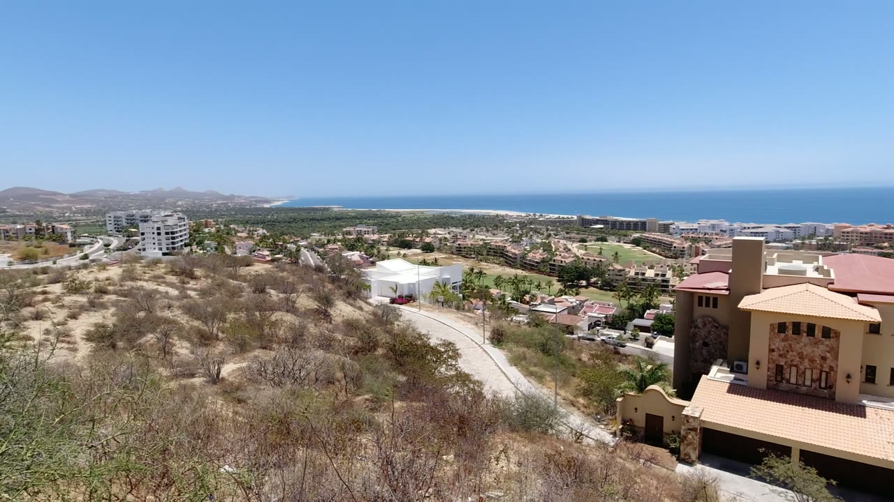 Wide elevated view in San Jose del Cabo, Mexico, with dry hillside brush in front, tan homes and roads below, inland mountains to the left, blue ocean on the horizon, and no visible people.