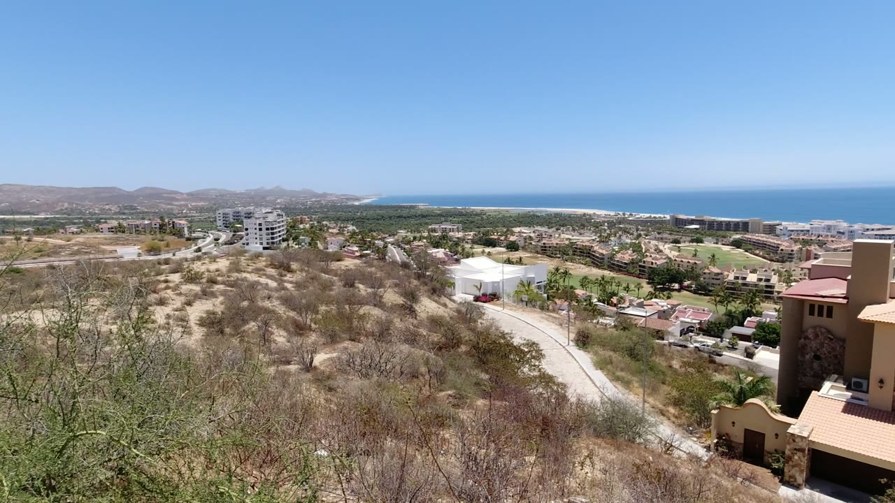 San Jose del Cabo hillside frame with brush and a tan roof at the right edge, residential streets and buildings below, green strips of landscaping, low mountains, ocean, and no visible people.