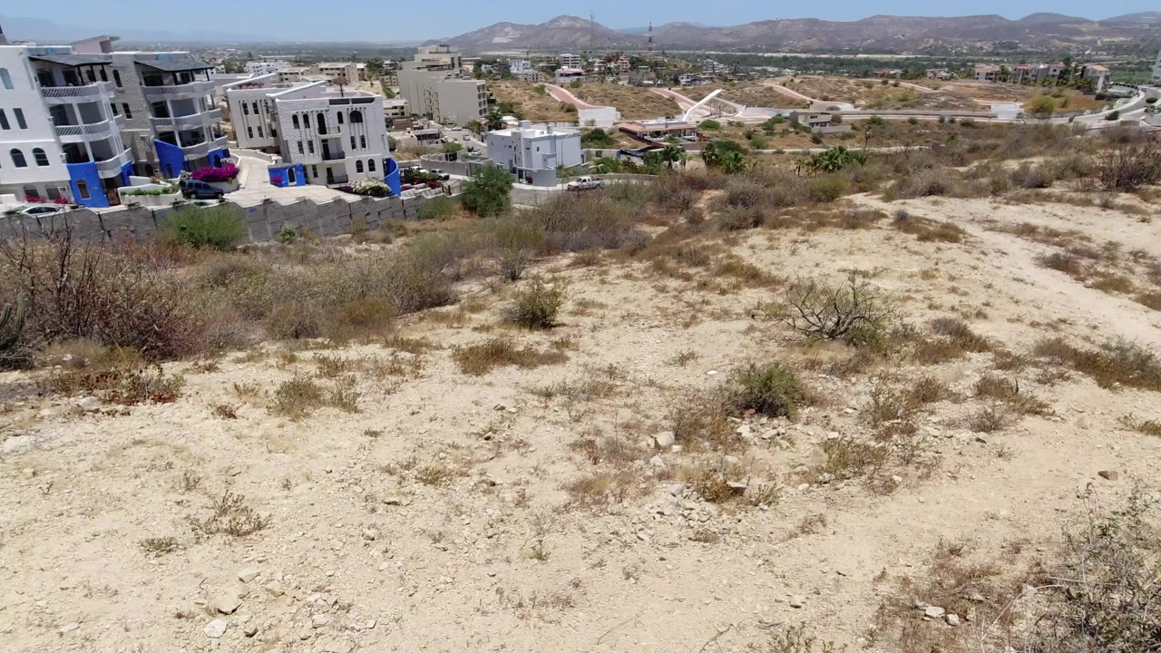Dry desert hillside with scrub, nearby buildings, distant hills, and blue sky.