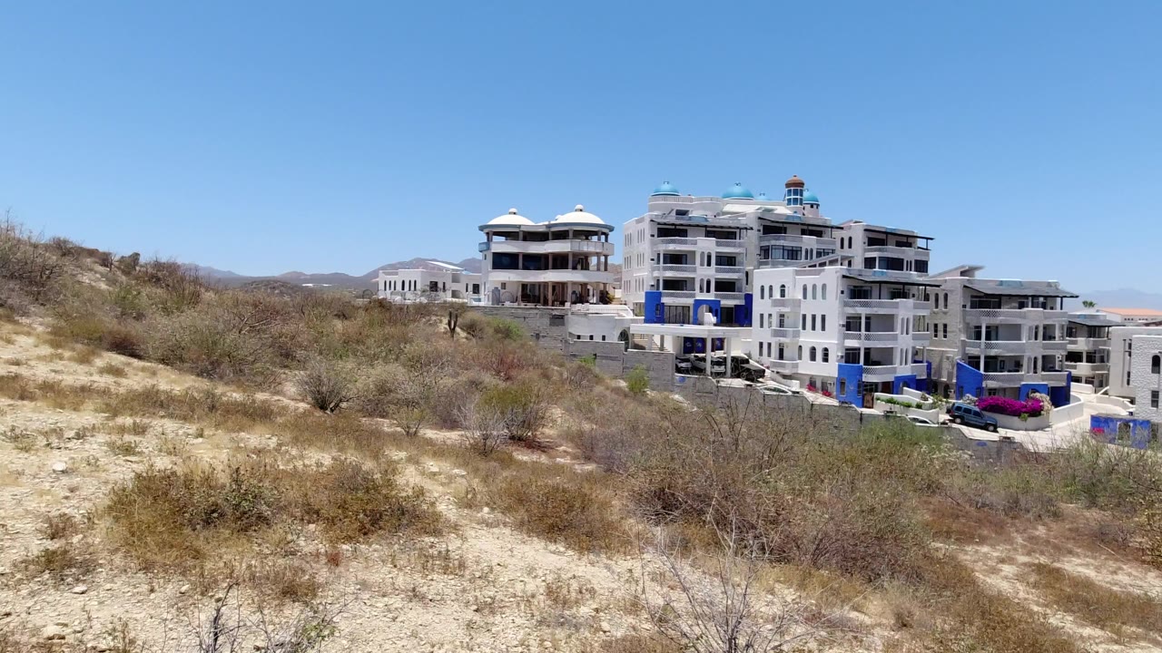 Grey and white buildings beyond dry hillside brush under blue sky.