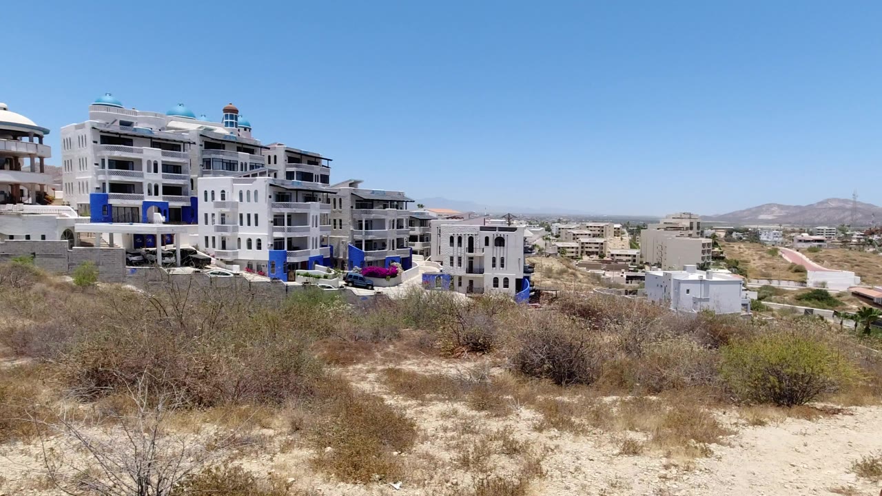 Wide elevated view of white hillside buildings, dry scrub, roads, distant hills, and blue sky.