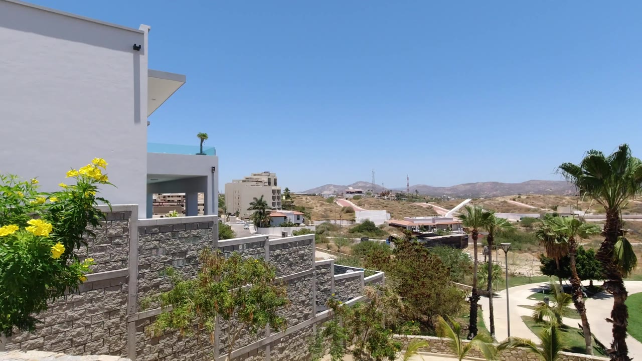 San Jose del Cabo frame looking past the left edge of a white building and layered stone walls toward palms, low hills, scattered structures, and no visible people.